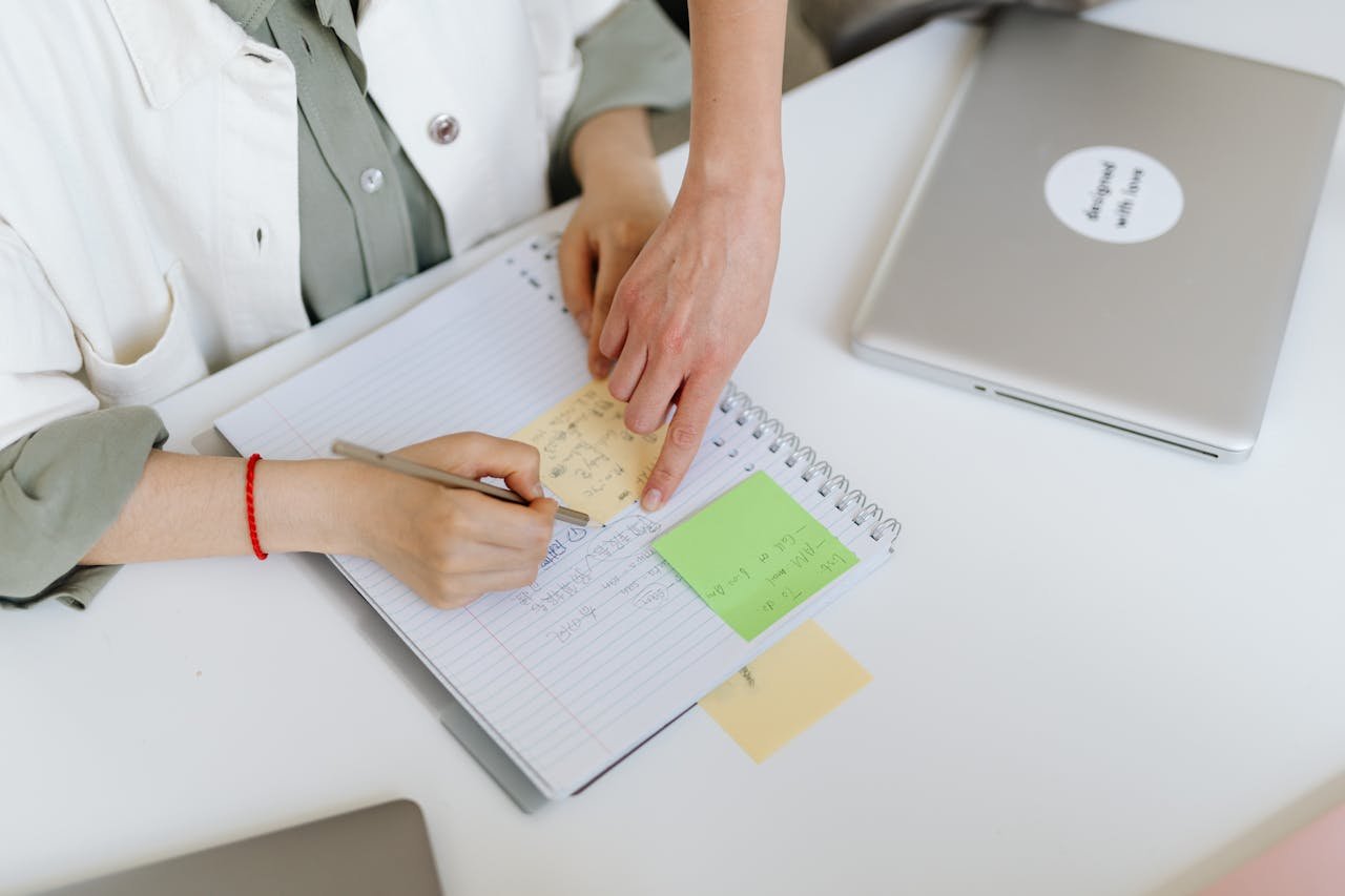 Two people collaborating over a notebook with post-it notes and a laptop in an office setting.